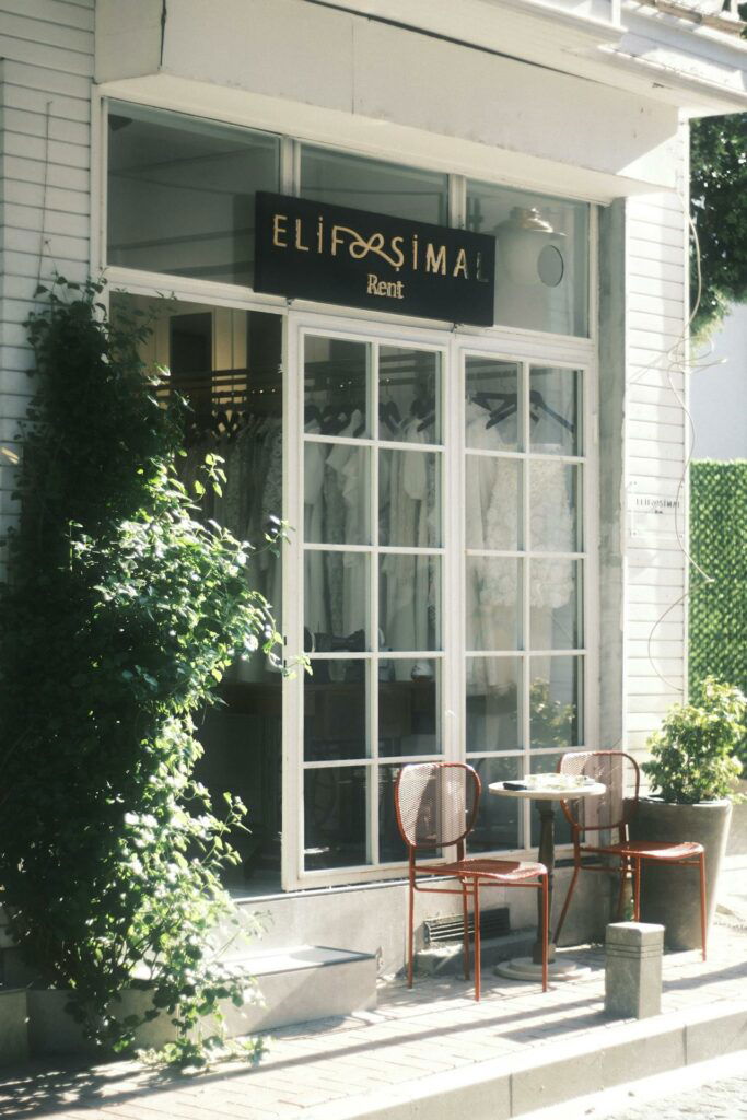 Storefront with large windows displaying clothing inside. Sign above reads "ELIF SIMAL Rent." Two red chairs and a small table are set outside, beside a potted plant and greenery.