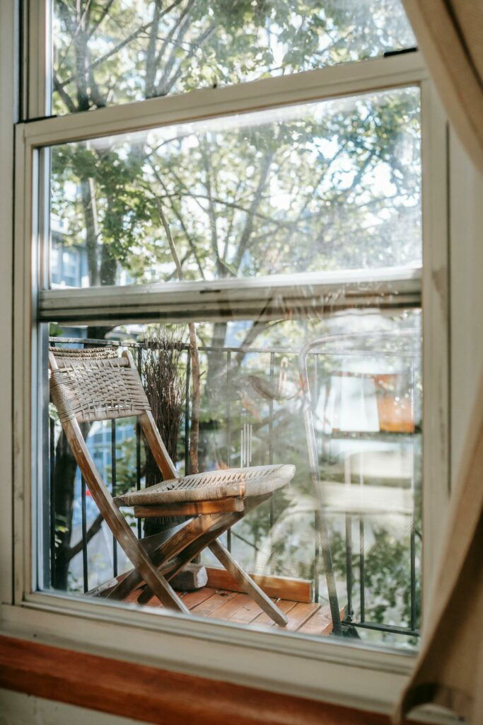A wooden chair is placed on a small balcony, viewed through a window with reflections of tree branches.