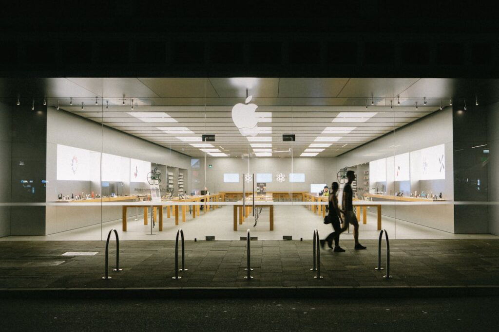 Exterior view of an Apple Store at night, featuring large glass windows, illuminated inside, with visible display tables and products. Two people walk past on the sidewalk in the foreground.