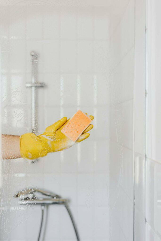 A person wearing yellow gloves is cleaning a glass shower door with an orange sponge in a white-tiled bathroom.