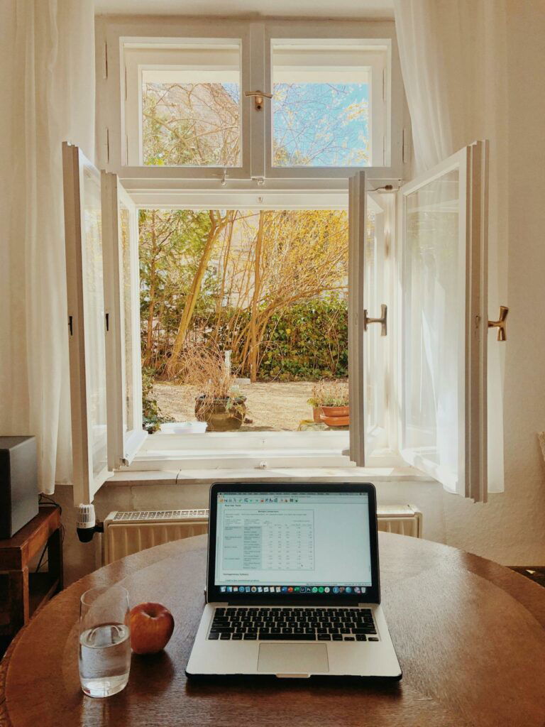 A wooden table with a laptop, a glass of water, and an apple is set near an open window, overlooking a garden with trees and plants. Bright daylight illuminates the room.