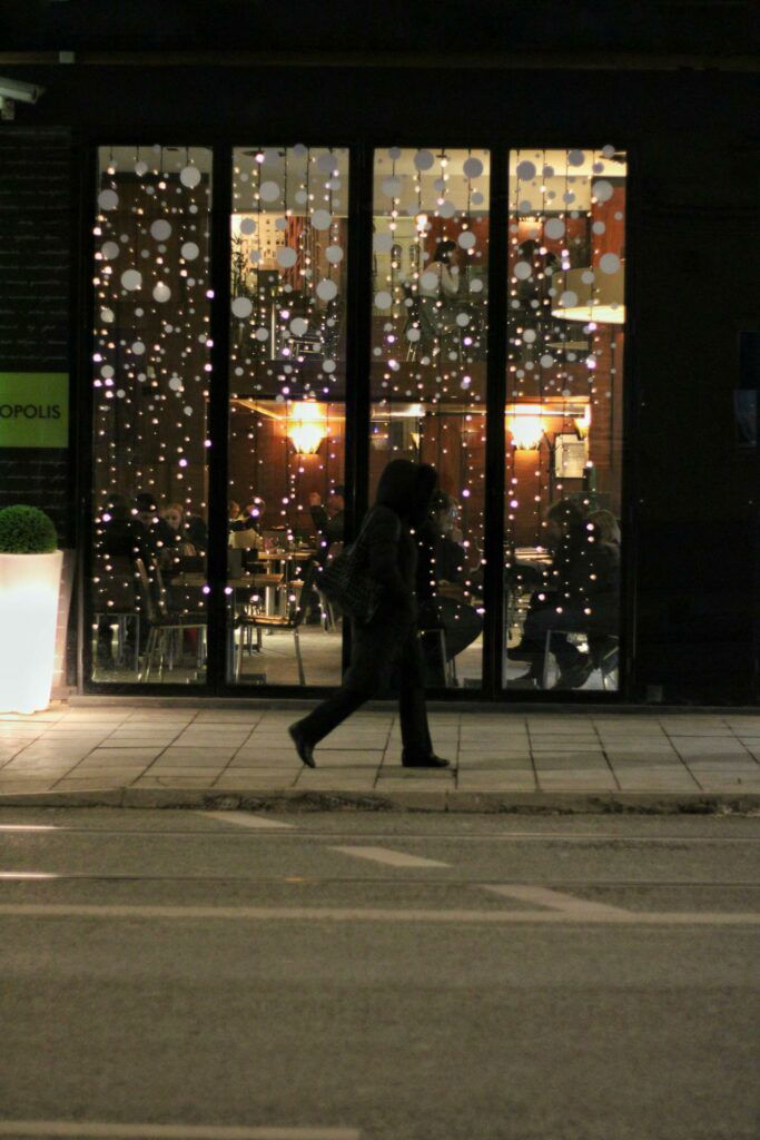 A person walks past a restaurant with large windows decorated with hanging spheres, at night. The interior is warmly lit and visible through the windows.