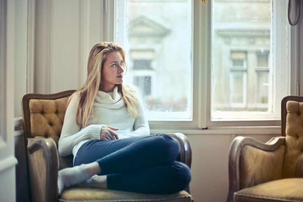 A woman in a white sweater and blue jeans sits on a cushioned chair inside a room, looking out of a large window.