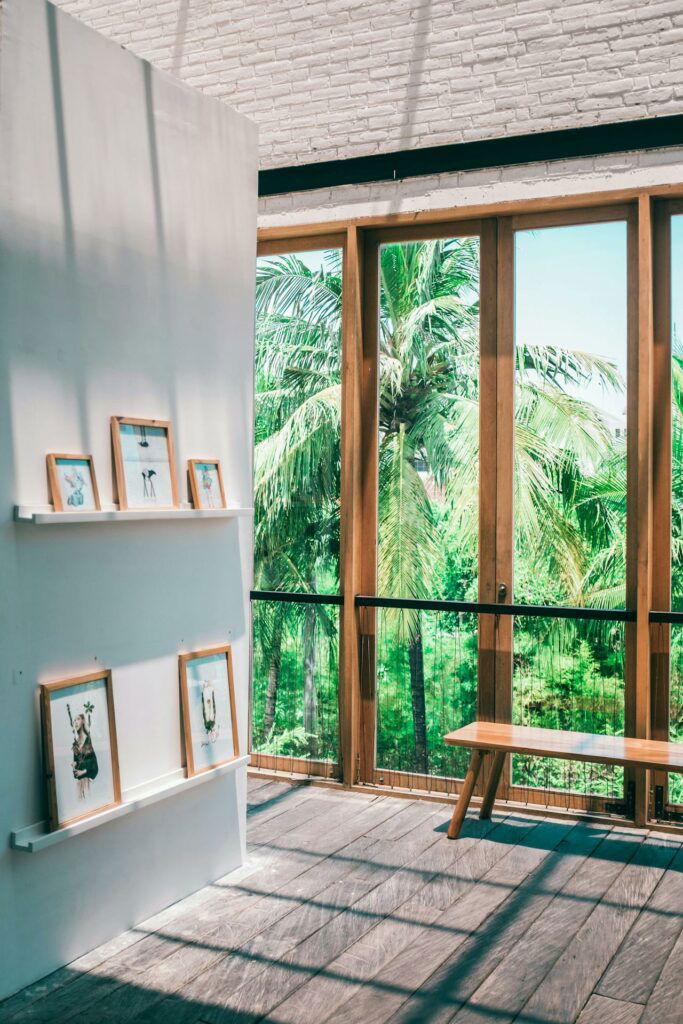 A sunlit room features framed pictures on white shelves and a wooden bench by large glass doors, with a view of lush palm trees outside.