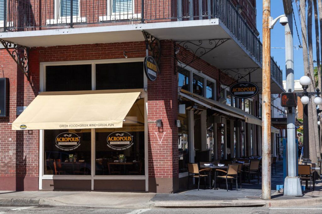 A corner brick building with a cafe named "Acropolis" featuring outdoor seating. The restaurant has signs and an awning advertising Greek cuisine, located in a sunny urban area.