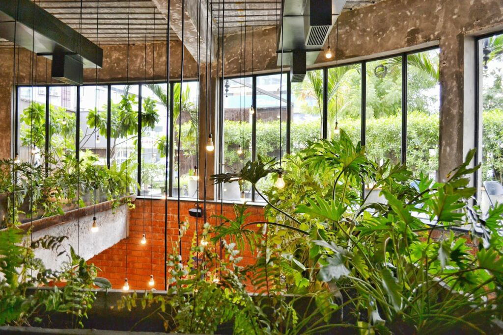 A modern indoor space with large windows, exposed brick walls, hanging light bulbs, and abundant greenery.